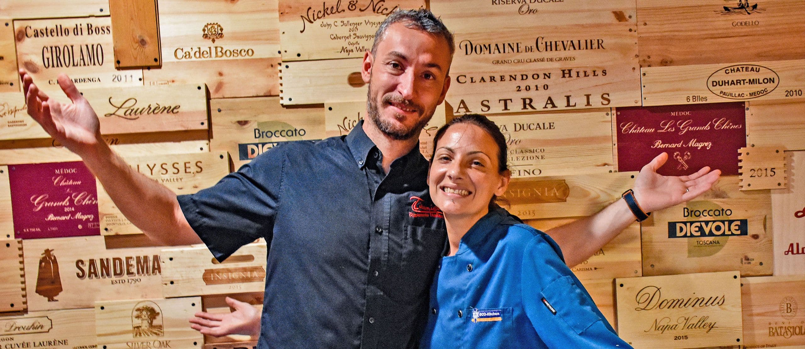 Luca and Monica welcoming guests in front of the restaurant's wine crate wall